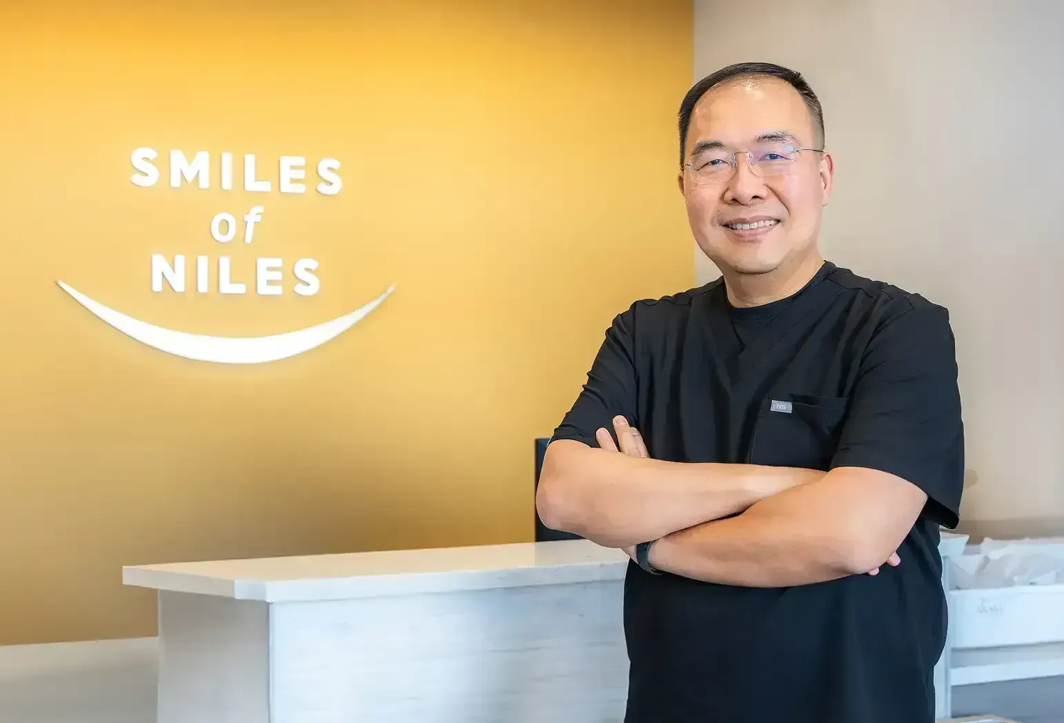 Smiling man in black scrubs standing with arms crossed by 