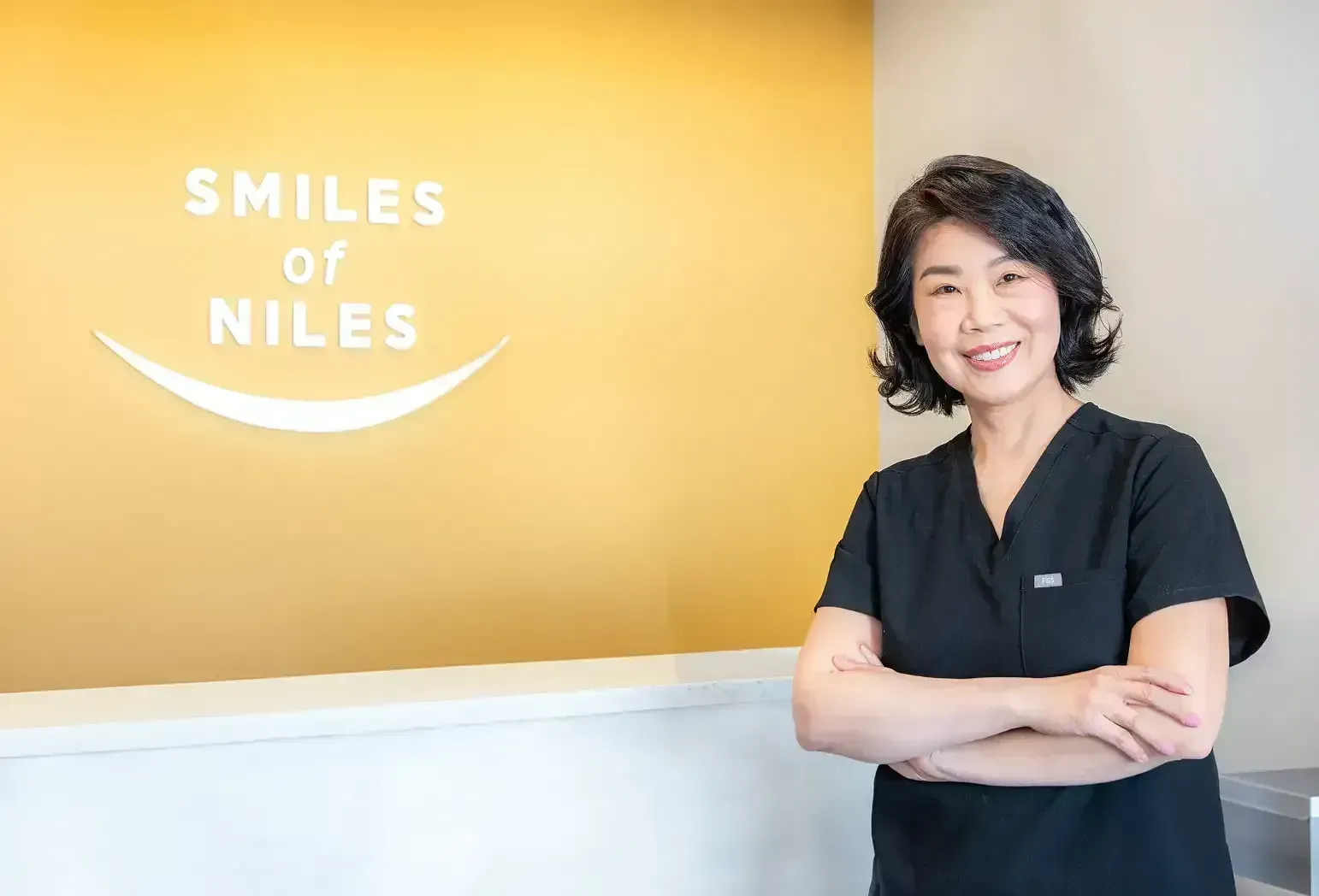 Smiling woman in black scrubs stands with arms crossed beside 
